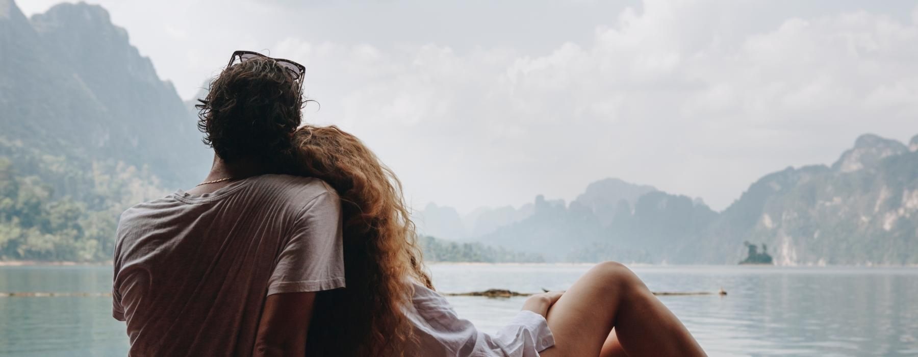 a man and woman sitting on a dock looking at the water and mountains